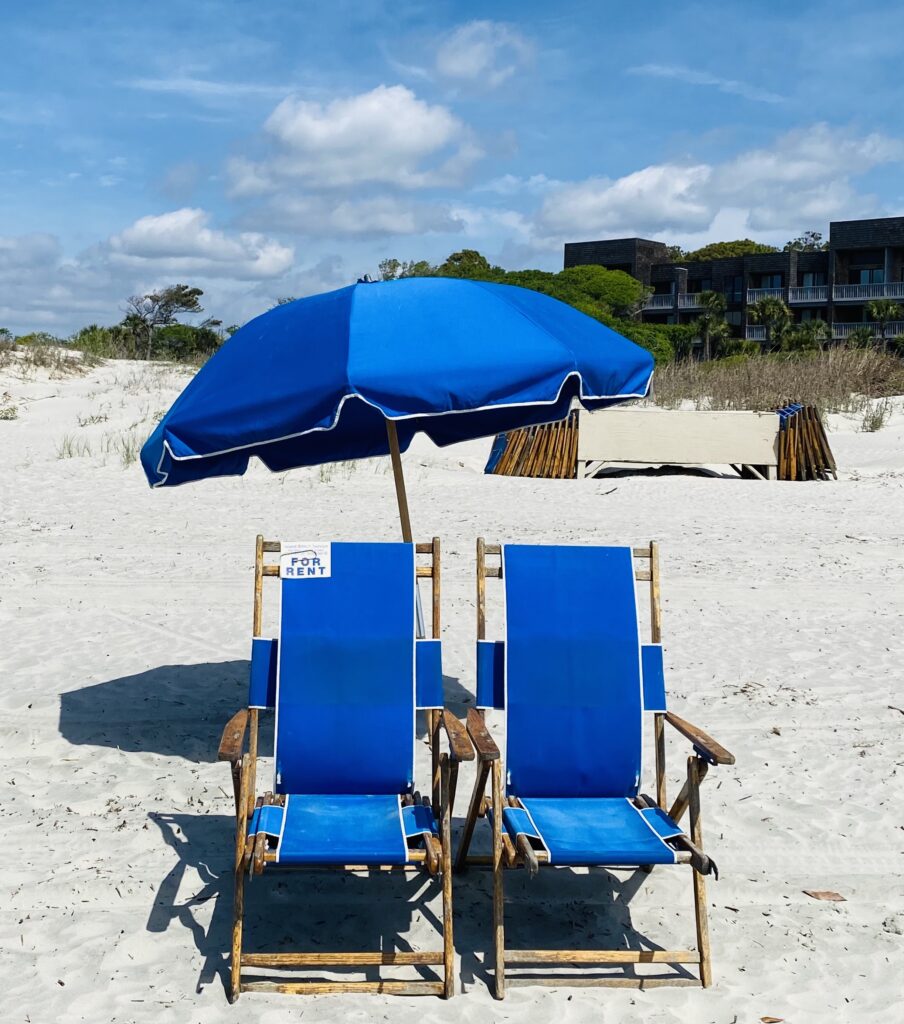 Two blue beach chairs and a large blue umbrella available for rent on the sandy shores of Kiawah Island.